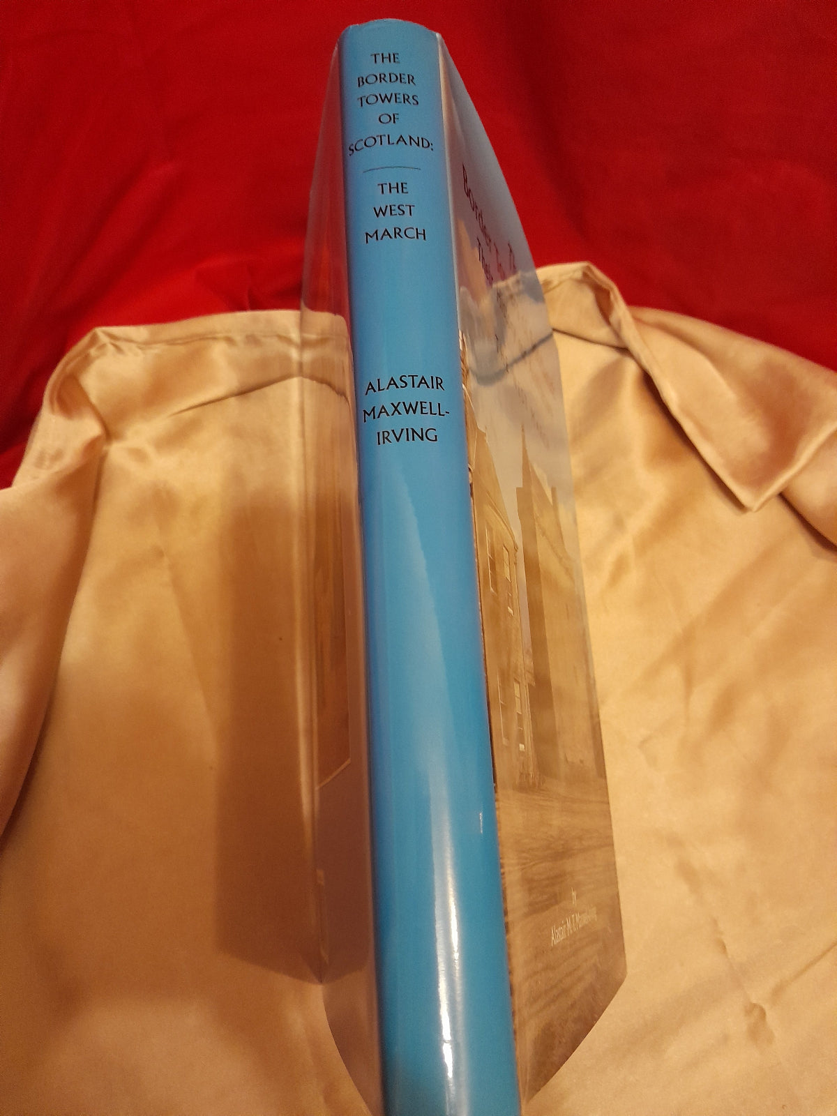 A hardcover book standing upright on a beige satin surface against a red fabric background. The spine is light blue with black text reading 'The Border Towns of Scotland', 'The West March', and 'Alastair Maxwell Irving'.