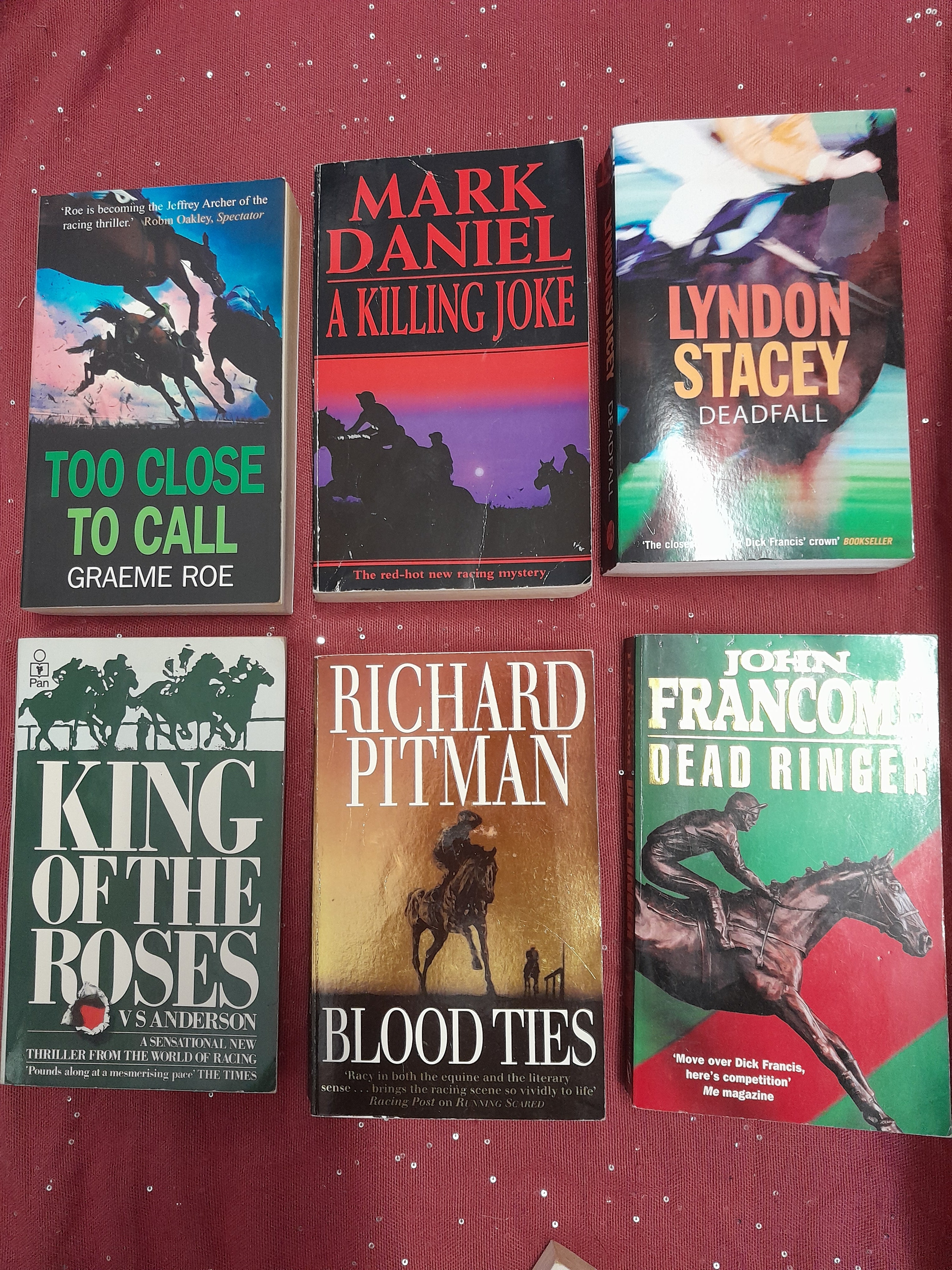 A collection of paperback books laid out on a deep red table cloth, with titles from the horse-racing crime fiction genre.