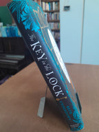 A hardcover book titled "The Key in the Lock" by Beth Underdown, propped open on a small stand on a wooden table. The book has a dark navy blue cover with teal floral illustrations and white lettering.
