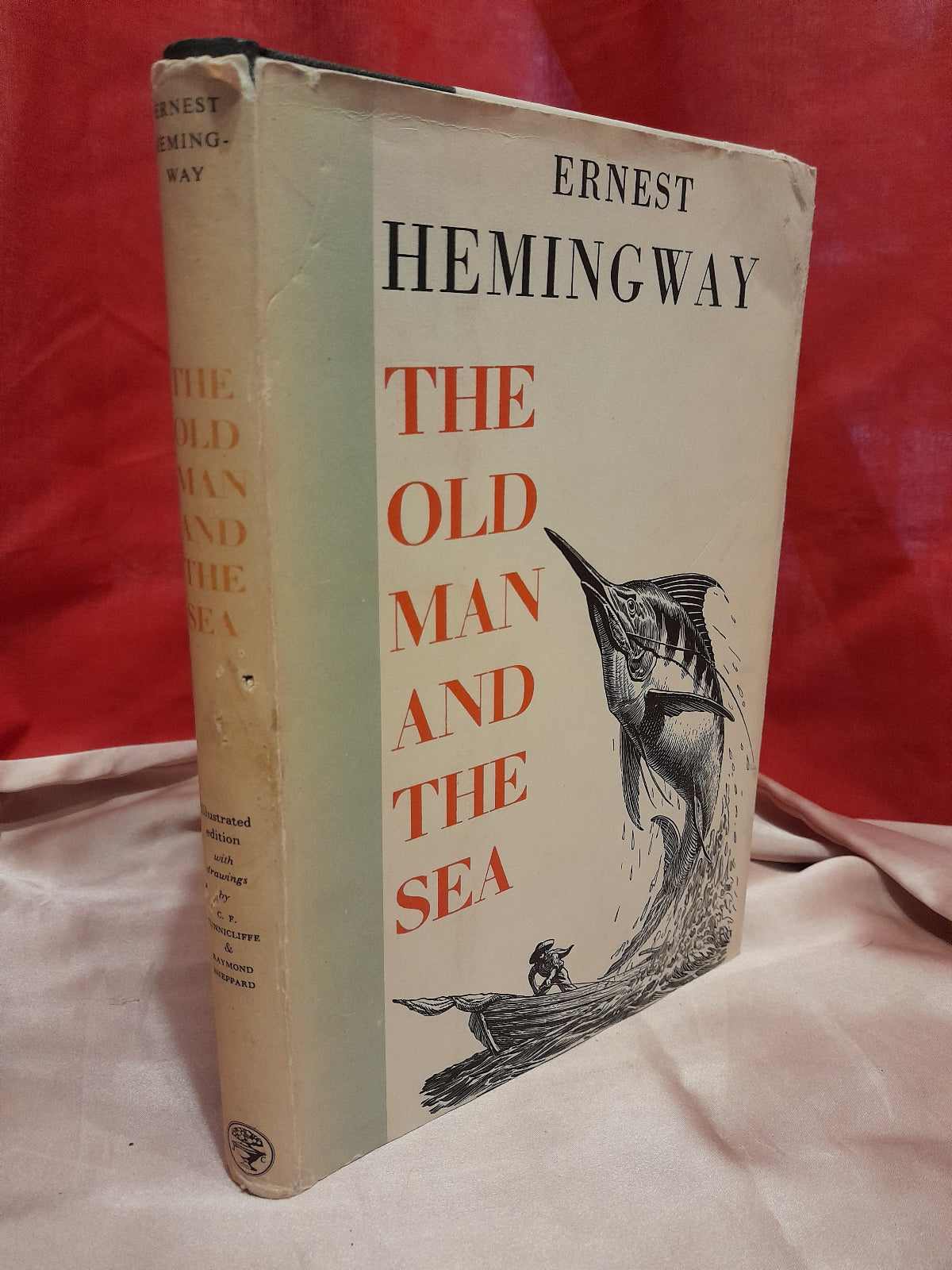Vintage hardcover book of Ernest Hemingway's 'The Old Man and the Sea' resting on a light-colored fabric surface against a red draped background.