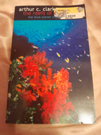 Arthur C. Clark's book 'The Reefs of the Pacific' showing a vibrant underwater coral reef scene with red and orange corals and small yellow fish, resting on a beige satin fabric background.